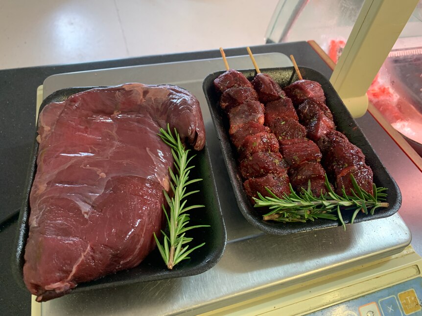 A piece of emu rump meat and three kebabs on skewers on trays on a butchers weigh scale. 