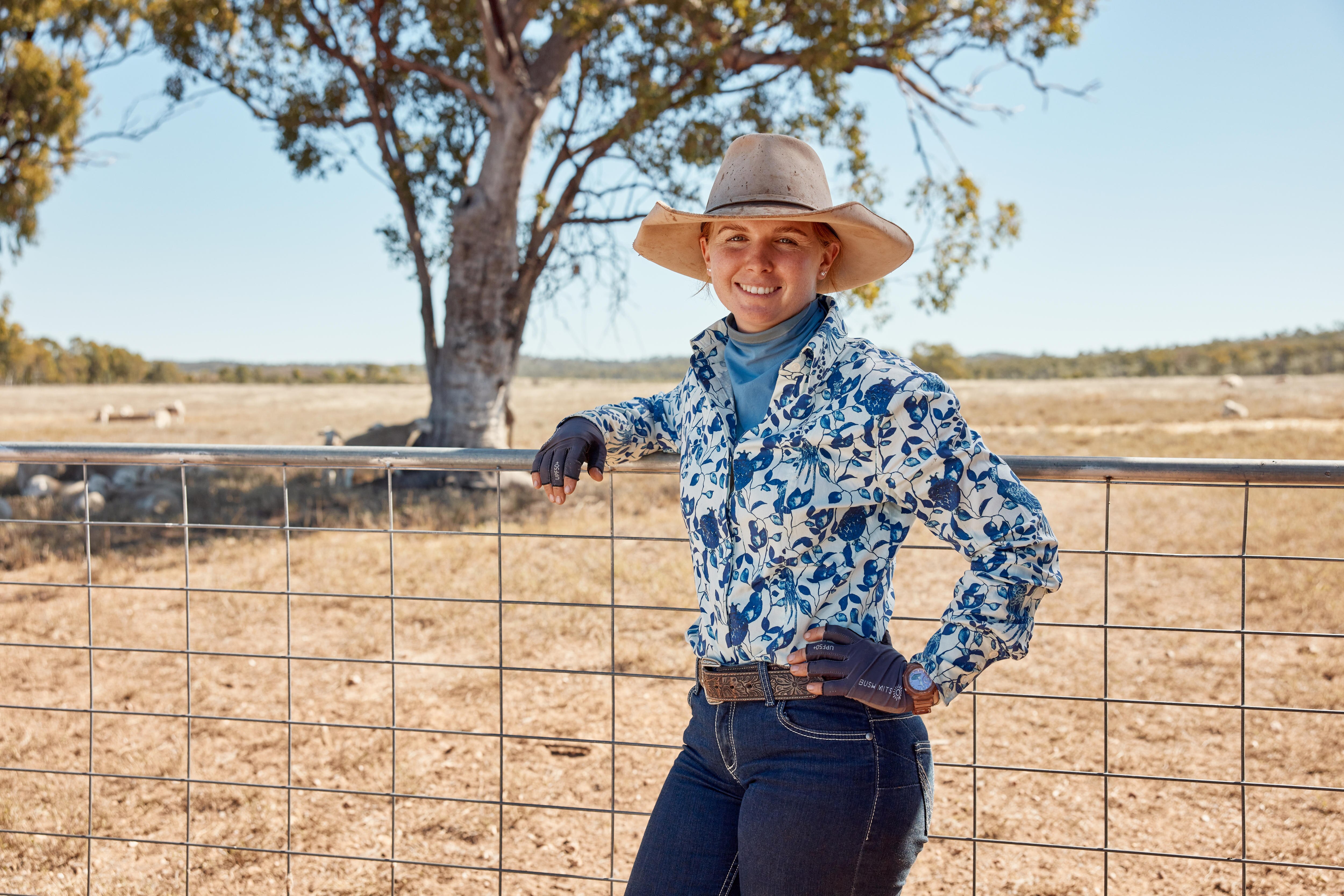 A woman wearing jeans, belt, collared shirt, fingerless gloves and a broad brim hat leans against a gate and smiles.