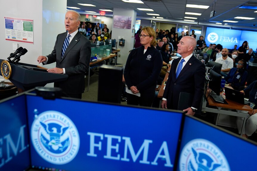 Un hombre blanco alto y anciano con traje habla desde un podio rodeado de funcionarios en una sala llena de trabajadores de FEMA frente a las computadoras.