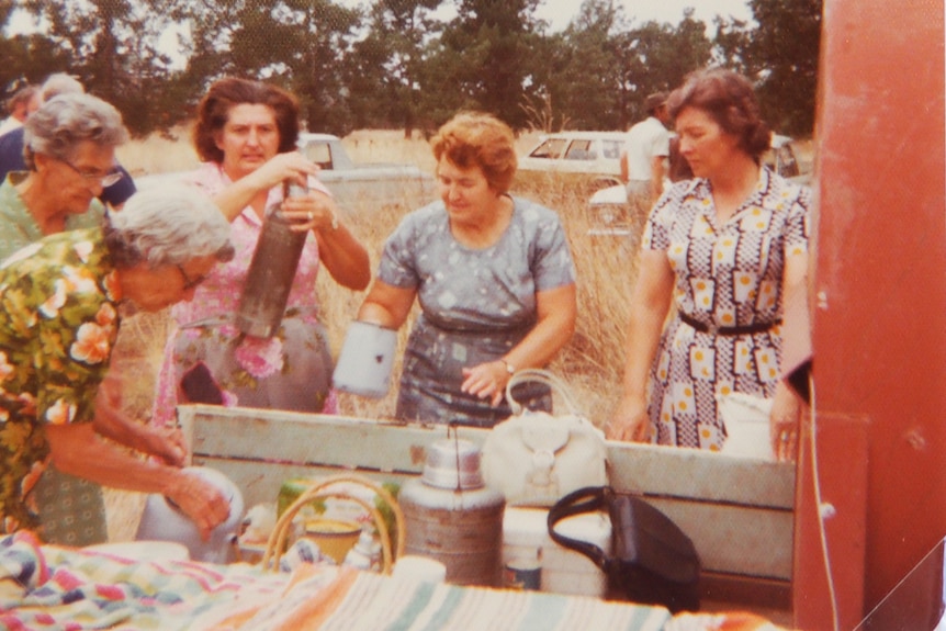 Five women in summer dresses huddle around the tray of ute full with baskets of food and a hot water urn.
