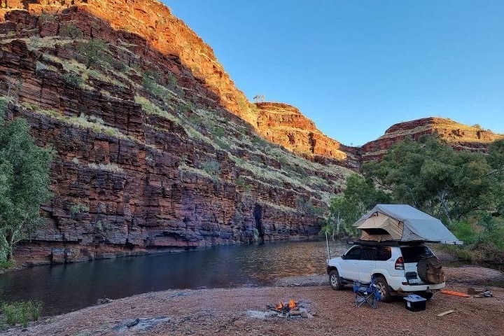 Une voiture, une tente et un feu de camp installés avec en toile de fond une gorge et de l'eau. 