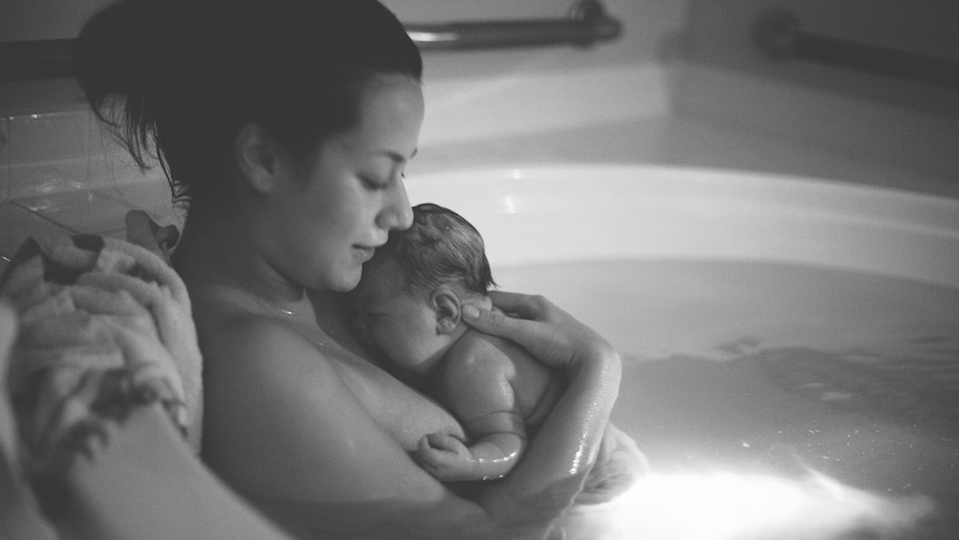 A mother holds her wet newborn to her chest as they sit in a birthing pool.