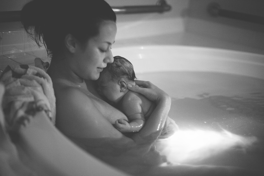 A mother holds her wet newborn to her chest as they sit in a birthing pool.