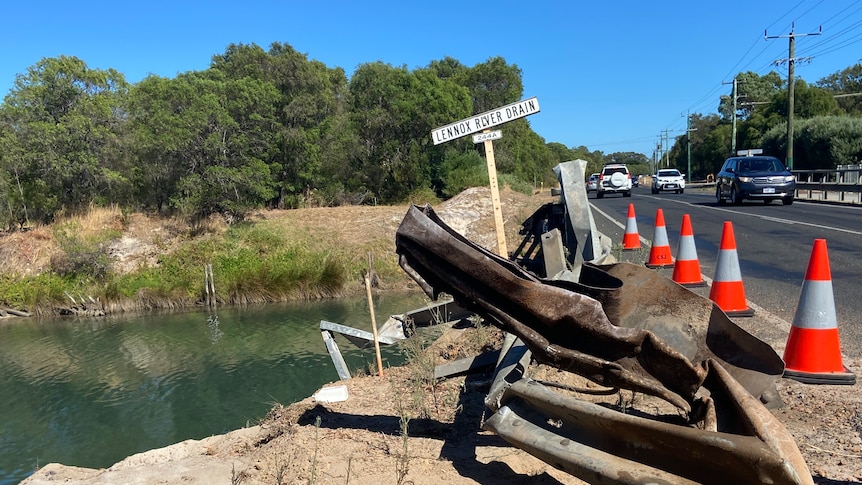 A road, with a bridge going over it, and a river. There's signs of damage to the bridge railing.