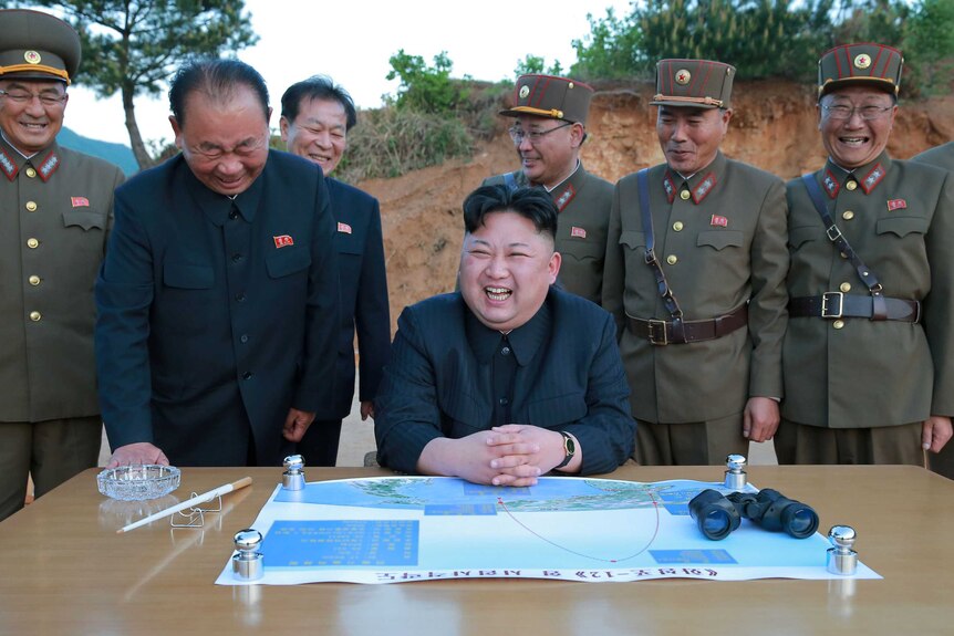 North Korean leader Kim Jong-un sits laughing in front of a map and a set of binoculars, surrounded by happy military members.