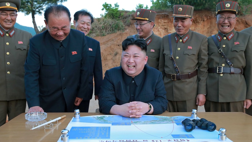 North Korean leader Kim Jong-un sits laughing in front of a map and a set of binoculars, surrounded by happy military members.