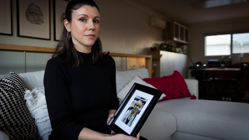 A woman wearing a dark dress sits on a couch and holds a framed photograph. 