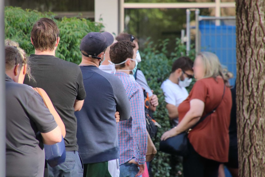 A queue of people, many wearing face masks, lines up in front of a hedge.