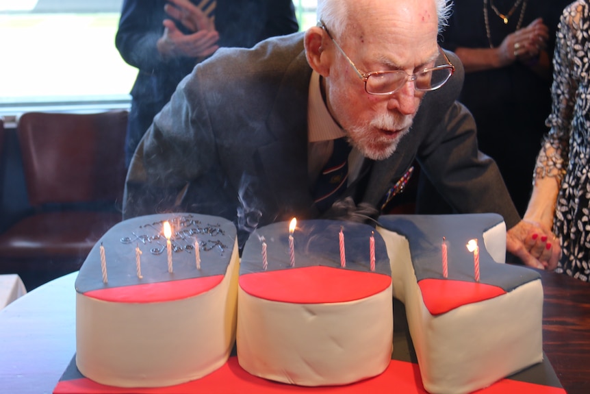 Bill Rudd bends down over a cake that spells out "100" and blows out his candles.