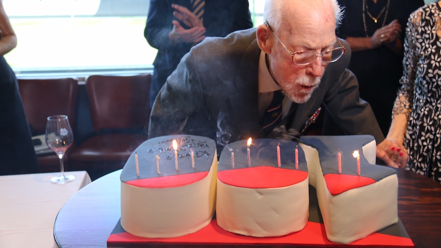 Bill Rudd bends down over a cake that spells out "100" and blows out his candles.