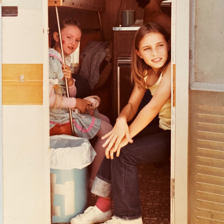 A young Claudia Karvan looks out of the door of a caravan or trailer, with another, younger girl just behind her.