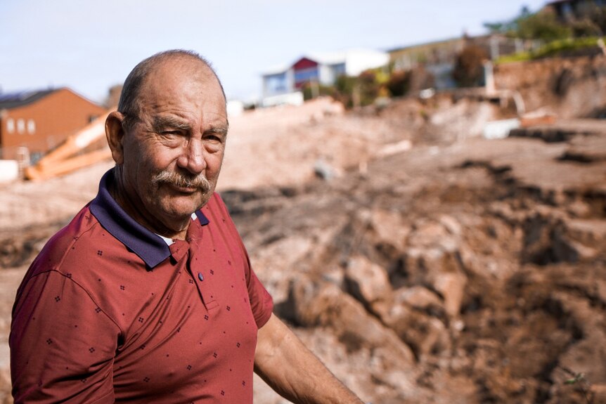 A man stands outdoors on the side of a hill with debris behind him