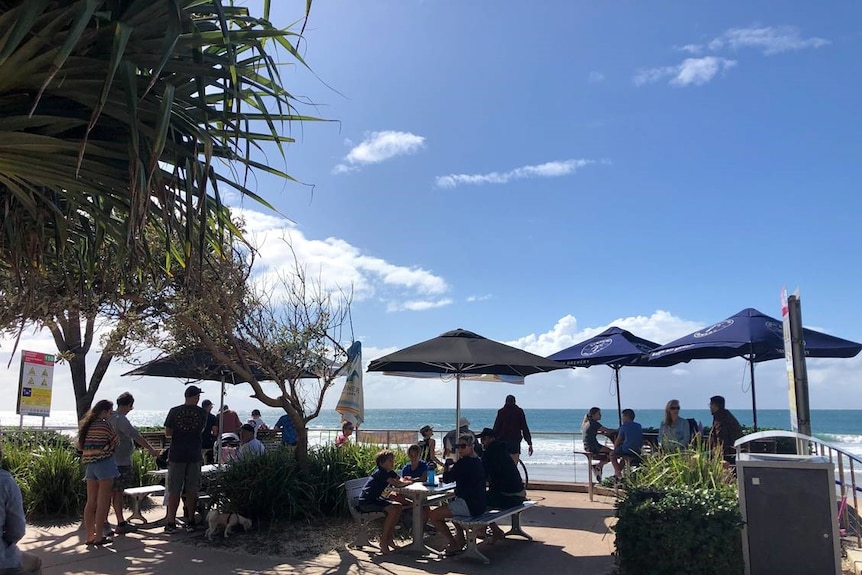 People crowd around outside tables at a beachside cafe and sit under large umbrellas on a sunny day