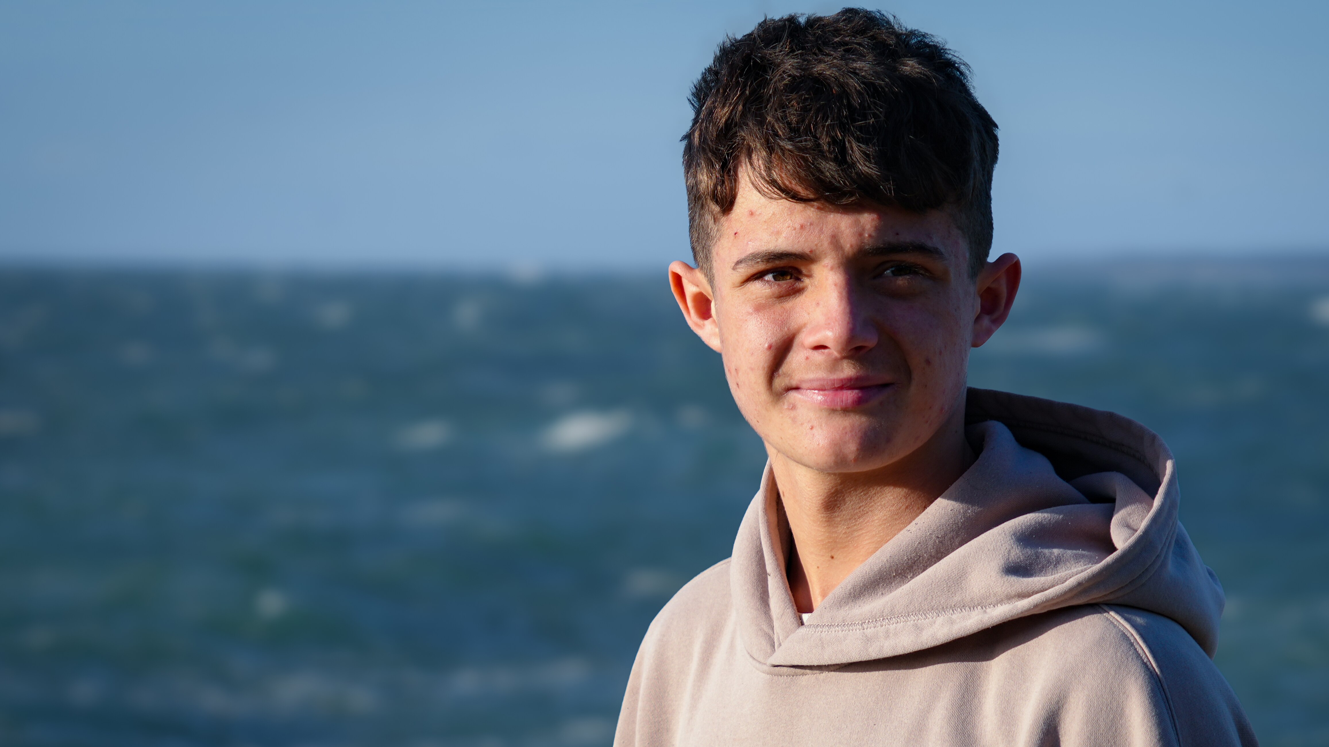 A young man in a grey hood smiles at the camera in front of an ocean with waves.