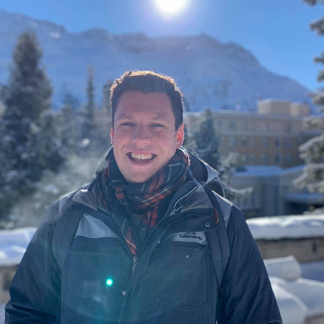 A man in winter coat and scarf in front of a mountain and building.