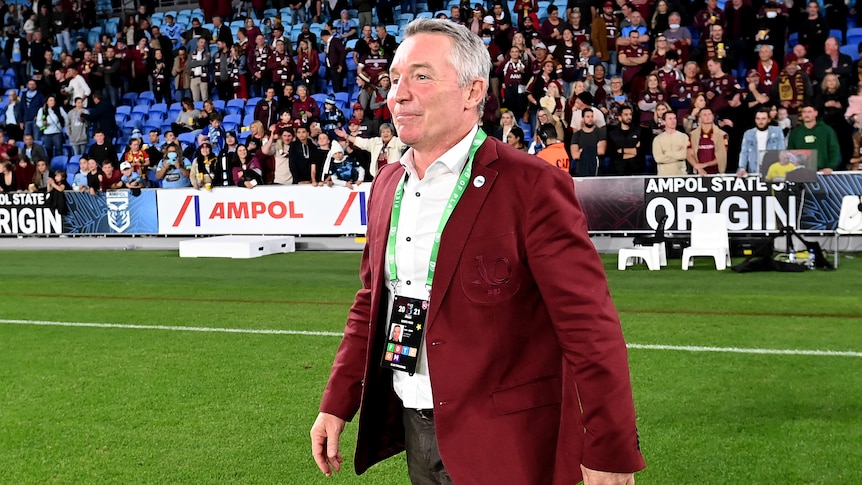 A coach wearing a Maroon jacket walks on the ground after a State of Origin game.