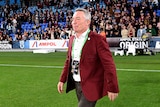 A coach wearing a Maroon jacket walks on the ground after a State of Origin game.