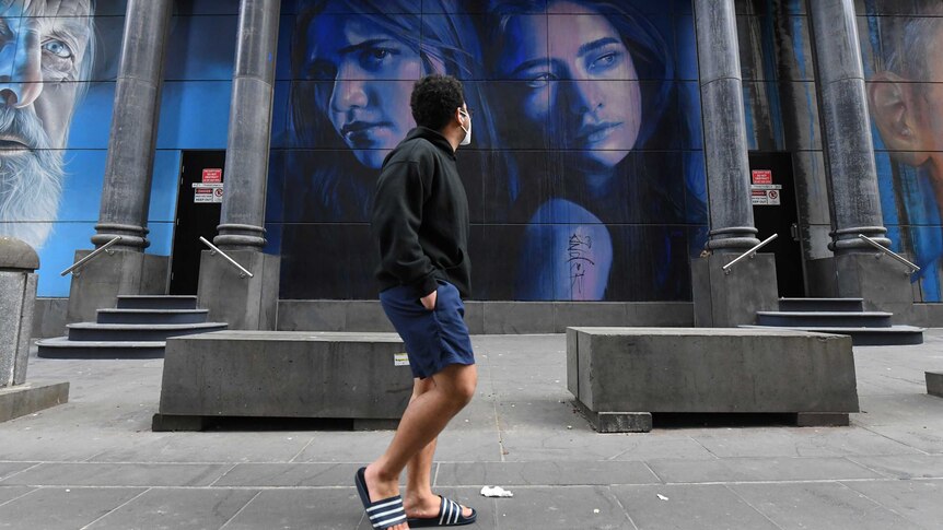 A young man wearing a face mask on a city street turns to look at painted mural of women's faces.