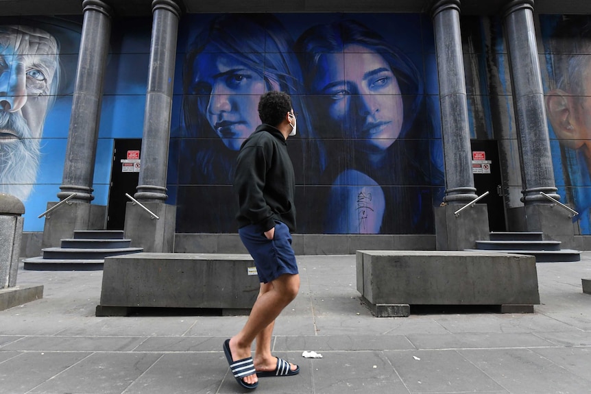 A young man wearing a face mask on a city street turns to look at painted mural of women's faces.