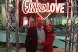 A couple stand under a neon sign that reads "Ekka Love". 