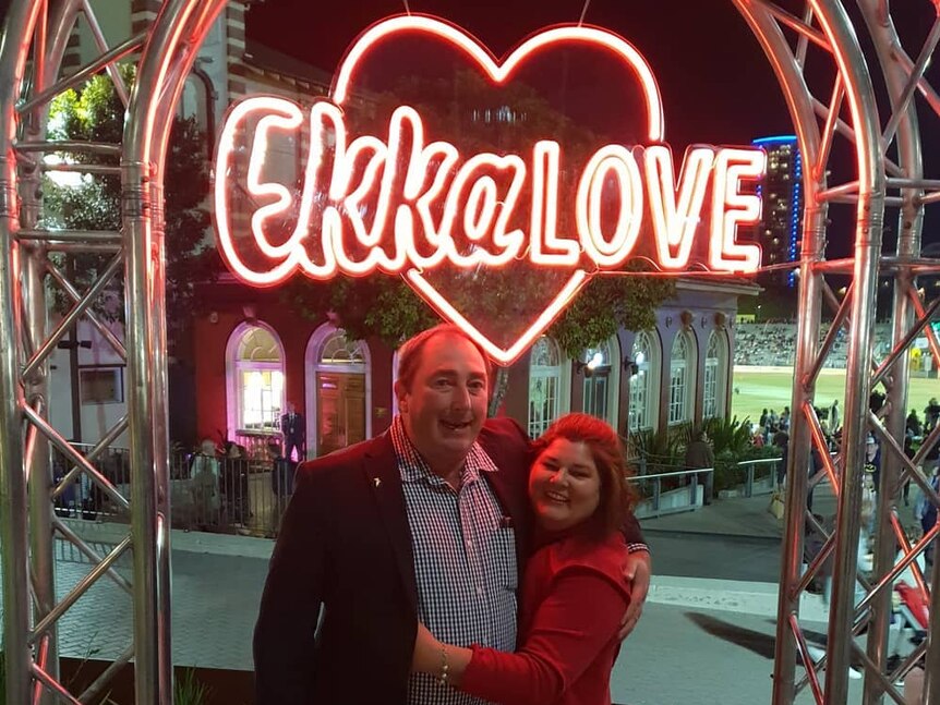 A couple stand under a neon sign that reads "Ekka Love". 
