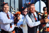 Two men play bagpipes during a march, with a drummer visible in the background