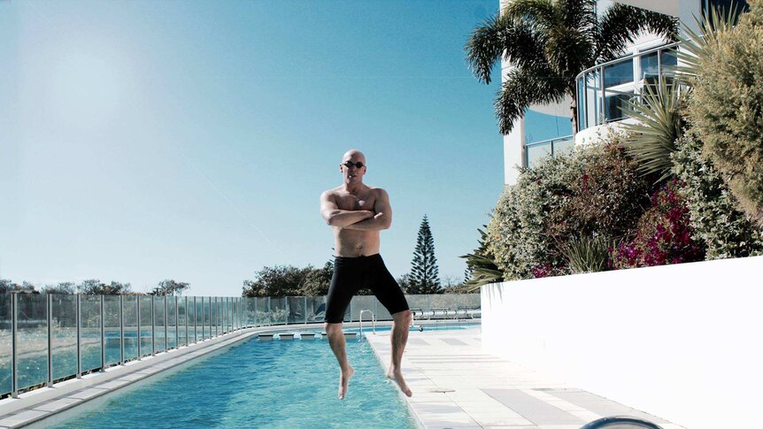 A man in swim shorts and goggles crosses his arms and strikes a mid-air pose as he jumps from the edge into a pool.