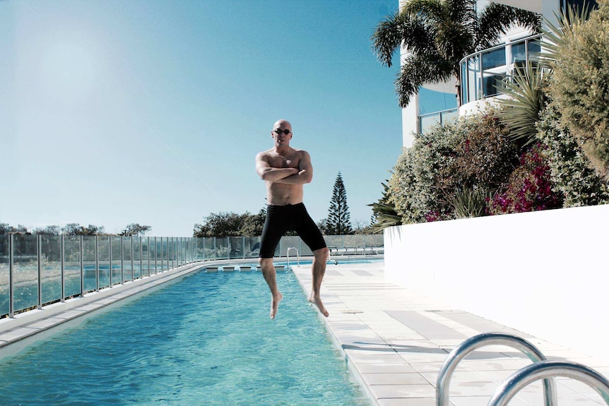 A man in swim shorts and goggles crosses his arms and strikes a mid-air pose as he jumps from the edge into a pool.