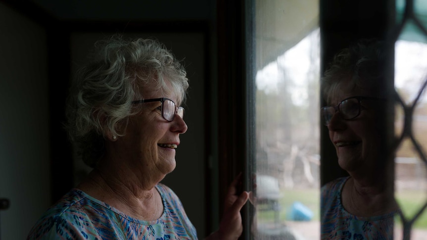 A woman stands near a window looking out, her face reflected back at her.