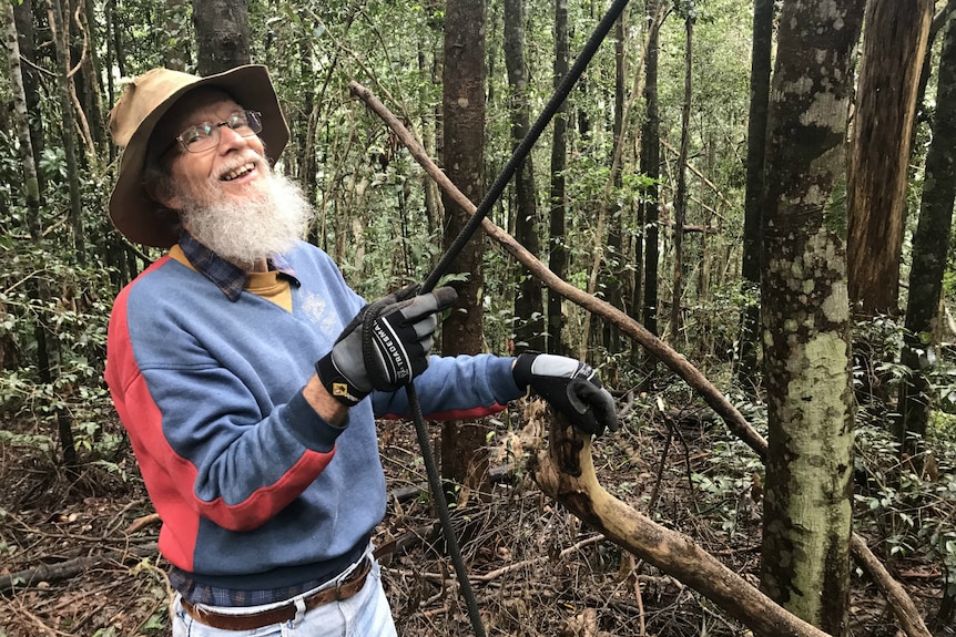 John Childs smiles at the camera as he holds a rope leading up into the trees.