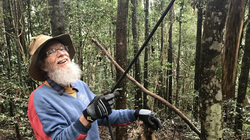 John Childs smiles at the camera as he holds a rope leading up into the trees.