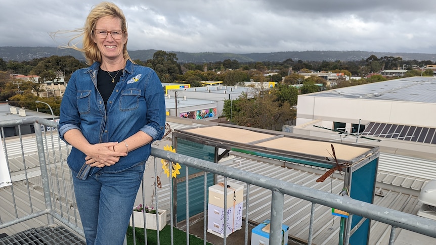 A blonde haired white woman Xandra Helbers is smiling and wearing a denim jumpsuit on a rooftop in front of hives.