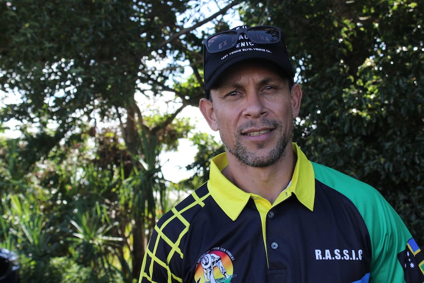 A man with sunglasses on his head wearing a green, yellow and black shirt smiles at the camera