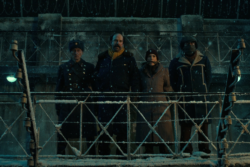 A guard, a man and two gagged captives stand on a platform in the snow, looking down.