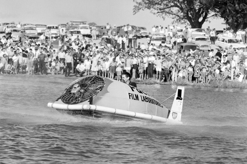 A black and white photograph of a hovercraft on a lake with onlookers.