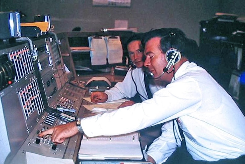 Two men wearing headsets sit at an operations console