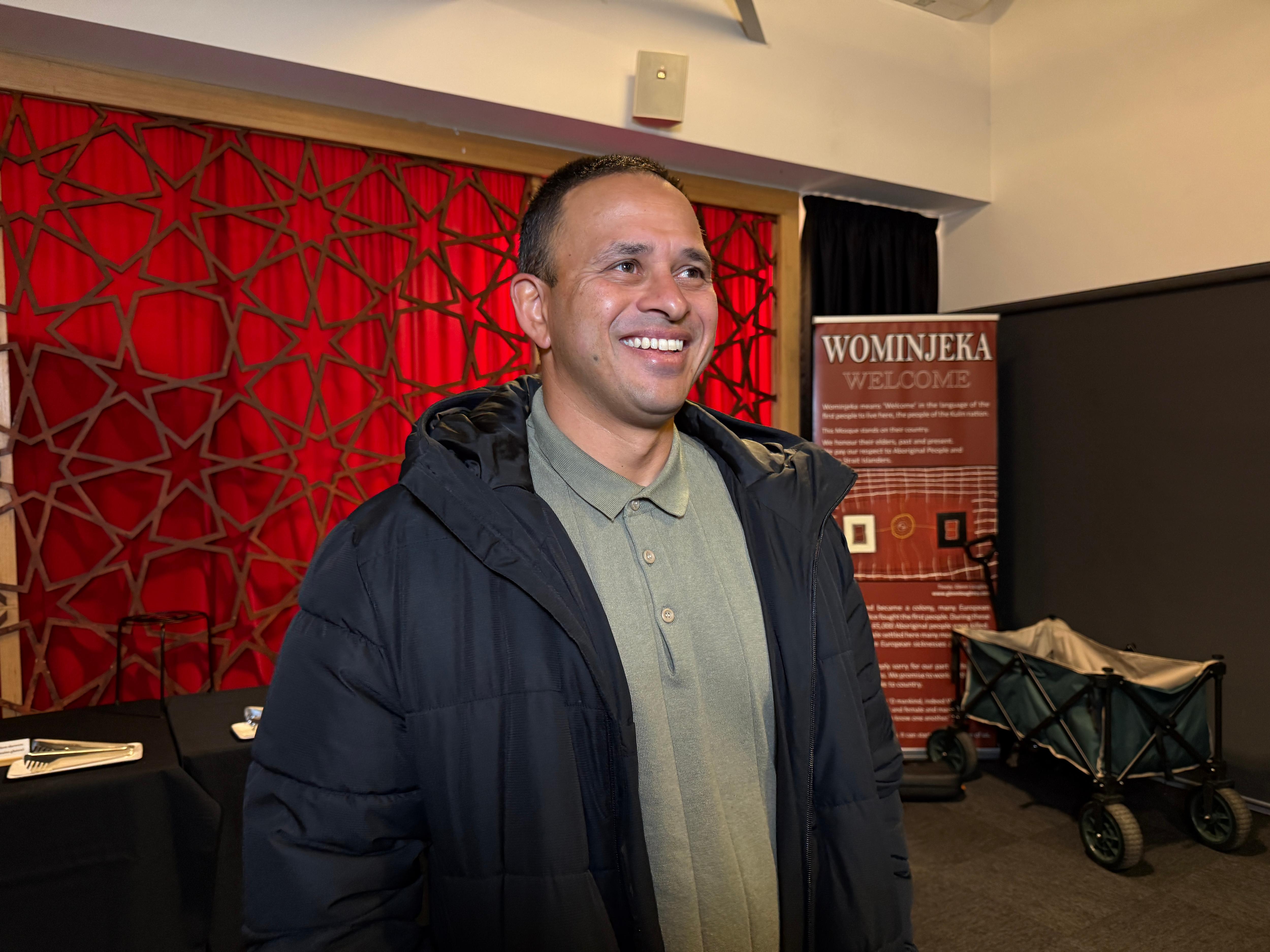 A middle-aged South Indian man in a green shirt and dark winter jacket smiles in front of red wall