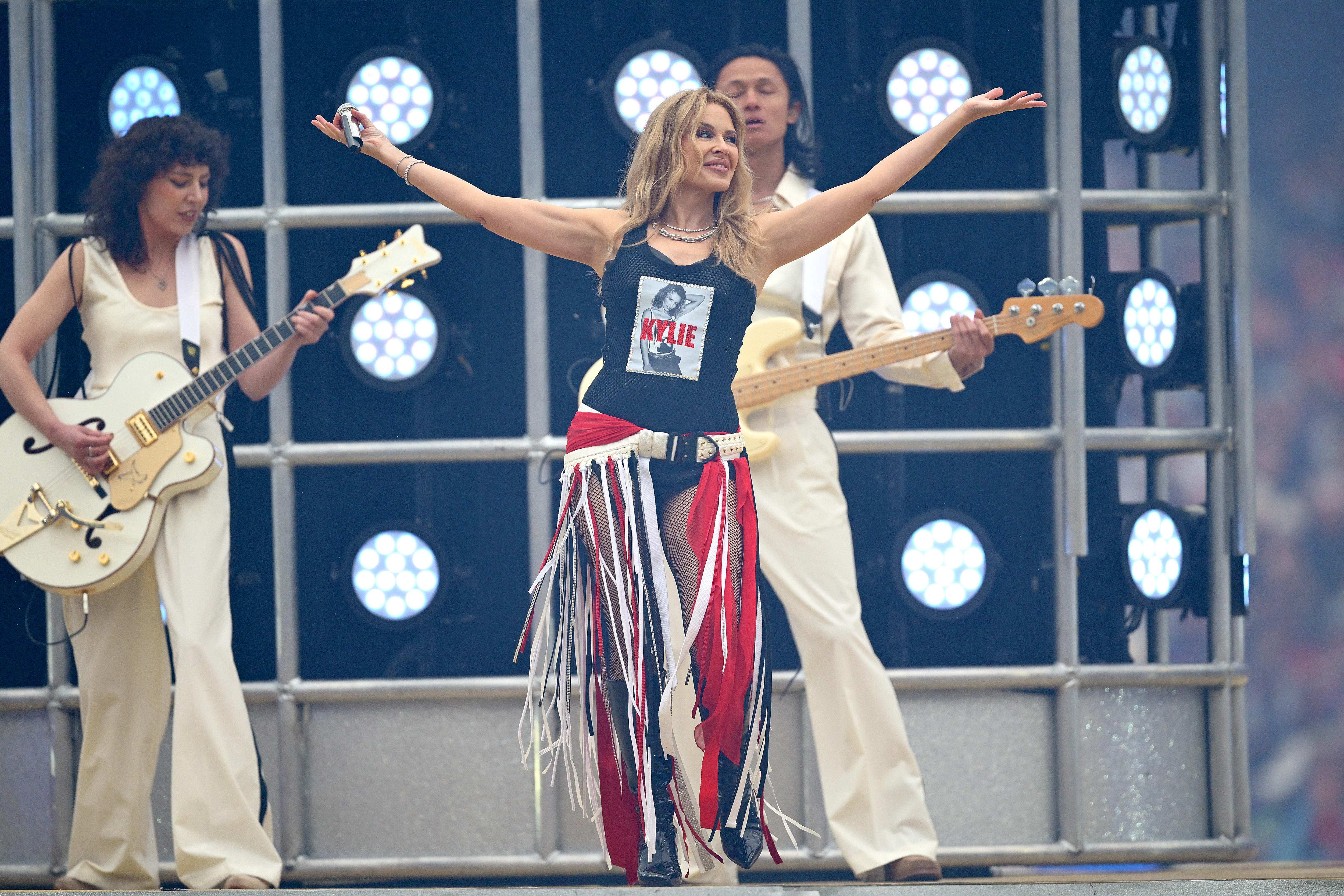Kylie Minogue wearing a black tank top with her face on it, and a white belt with red, white and black strips