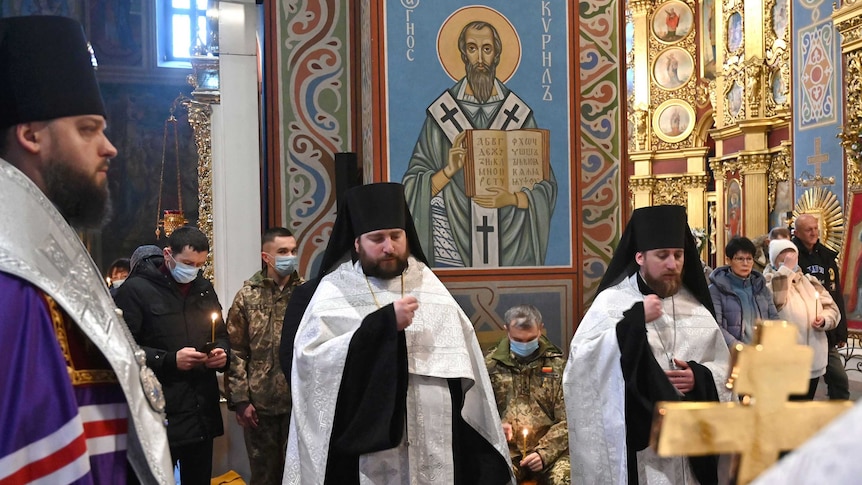 two ukranian orthodox leaders stand in front of civilians and soldiers in church hall ordained with orthodox iconography