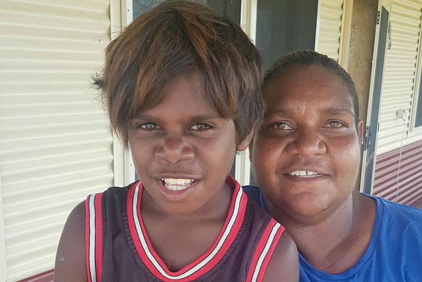 A young Indigenous boy standing next to an Indigenous woman, in front of a building with corrugated siding.