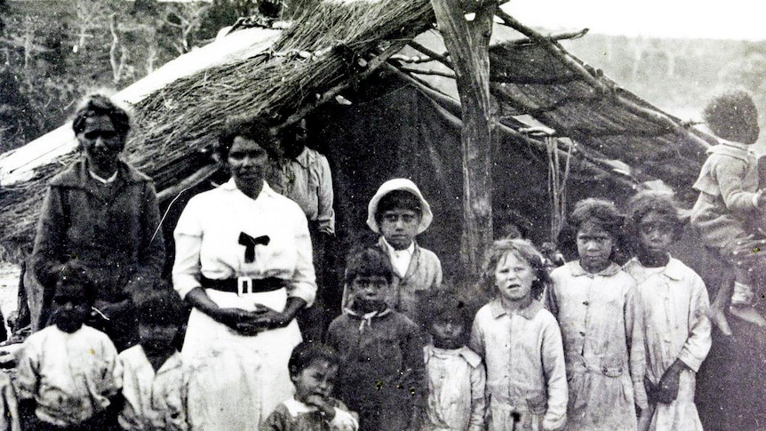 Two woman in black and white outfit stand with children in front of a shelter made of logs and twigs.