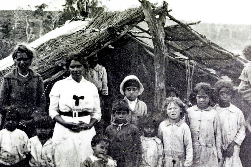 Two woman in black and white outfit stand with children in front of a shelter made of logs and twigs.
