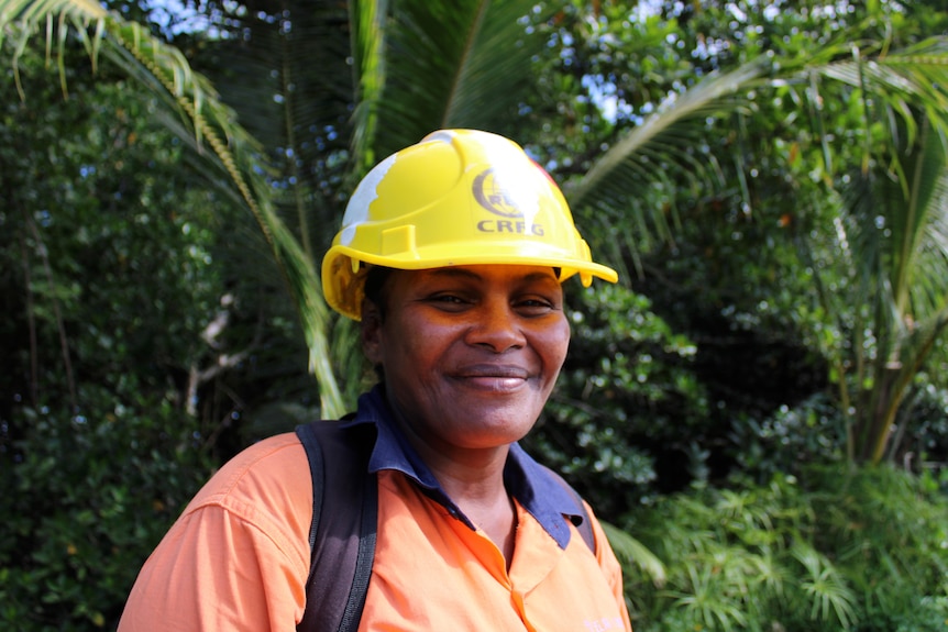 A woman smiling while wearing a yellow hard hat with tropical green trees in the background.