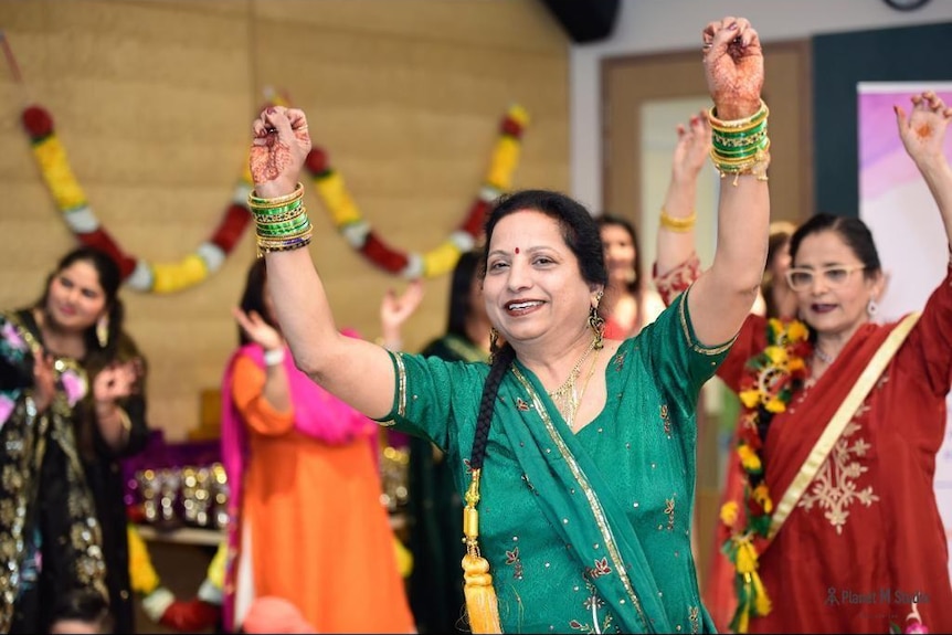 An Indian woman wearing green salwar kameez, hair braided, yellow ribbons at the end of pigtail, arms in air, dances with other.