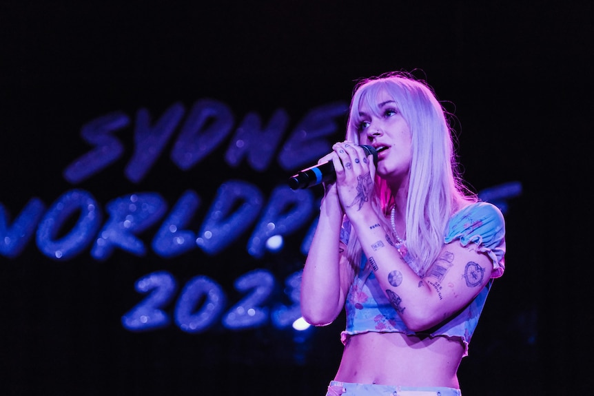 A woman sings into a microphone in front of a screen reading "Sydney WorldPride 2023"