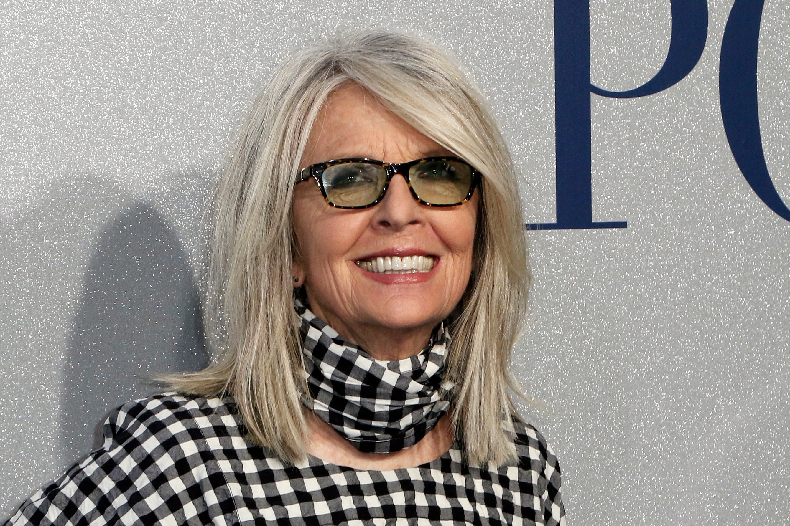 Diane Keaton, wearing tinted glasses and black and white chequed top and scarf, smiles in a head-and-shouloder shot