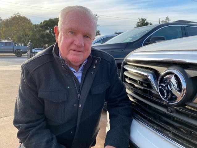 Holden dealer Neil Beer kneels down next to a car bonnet with the Holden logo in view.