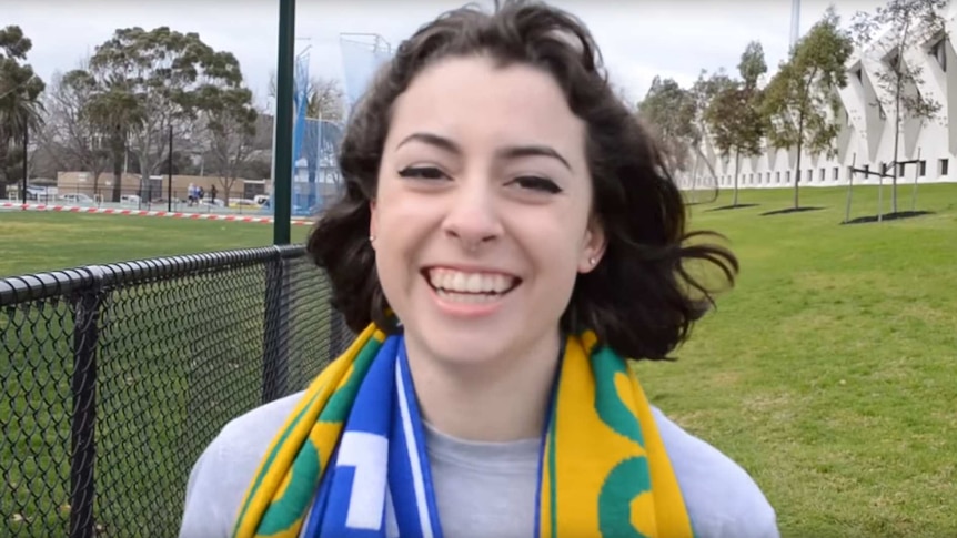 A young woman wears two football scarves, smiles at camera, football pitch in background.