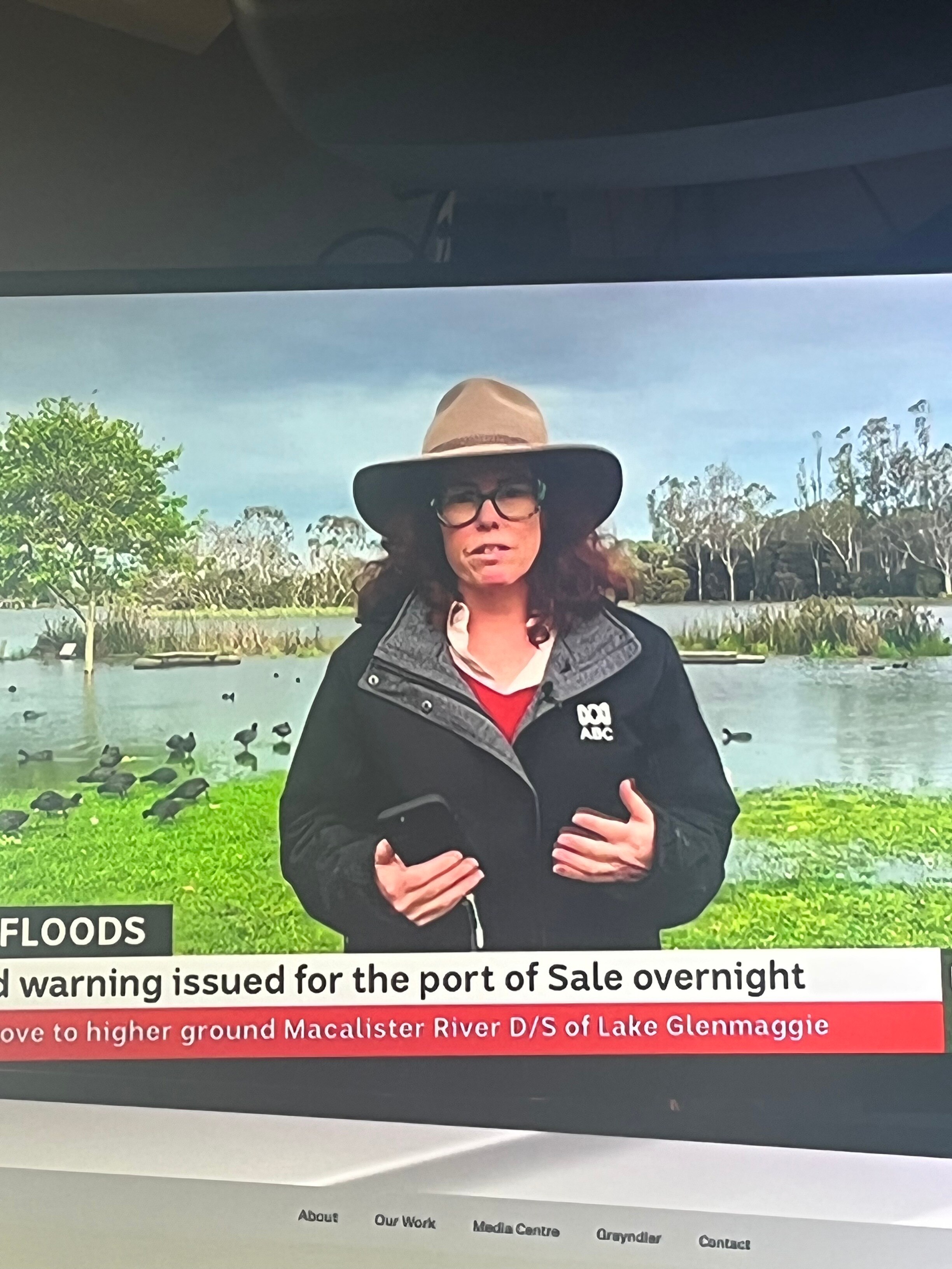Woman on TV screen, wearing an akubra hat, with floods in background.
