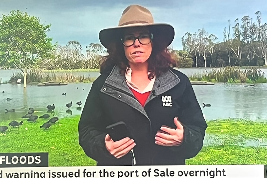 Woman on TV screen, wearing an akubra hat, with floods in background.